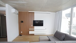 Una sala moderna con un sofá gris, un televisor de pantalla plana montado en una pared de paneles de madera, y un ventanal con vistas a la ciudad.