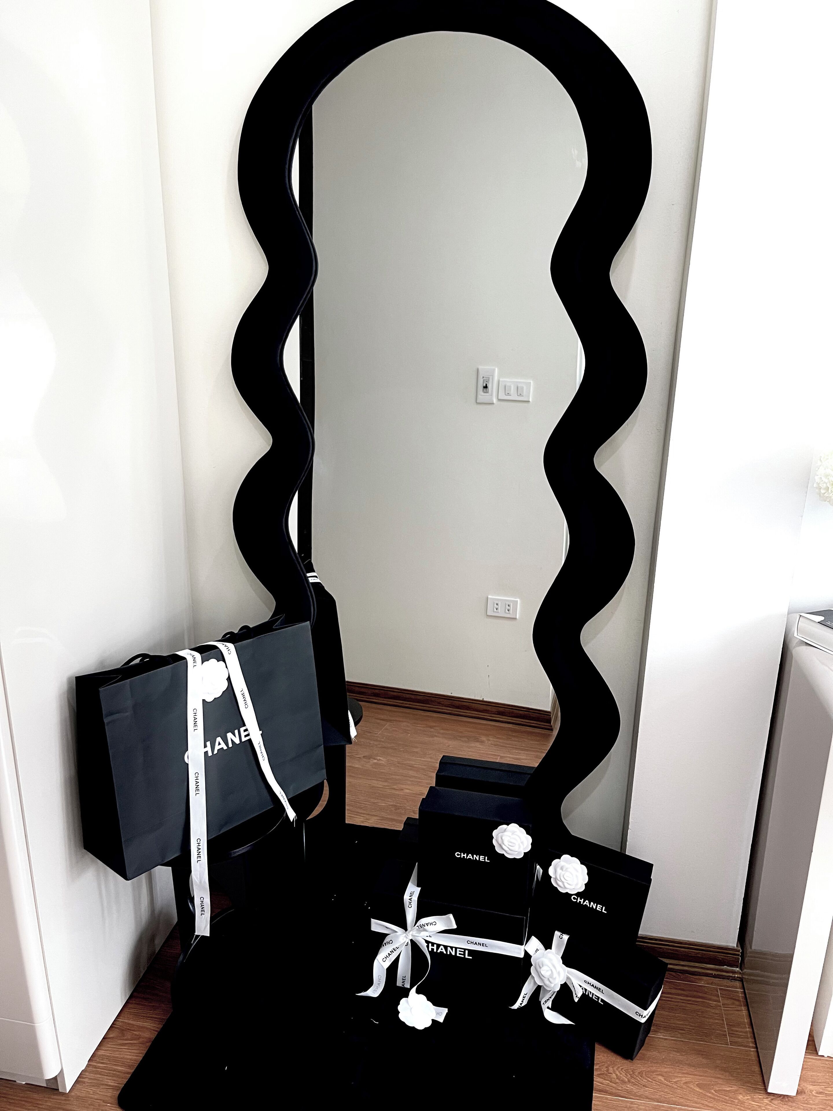A room with a unique black wavy mirror, a white wall, and a wooden floor.