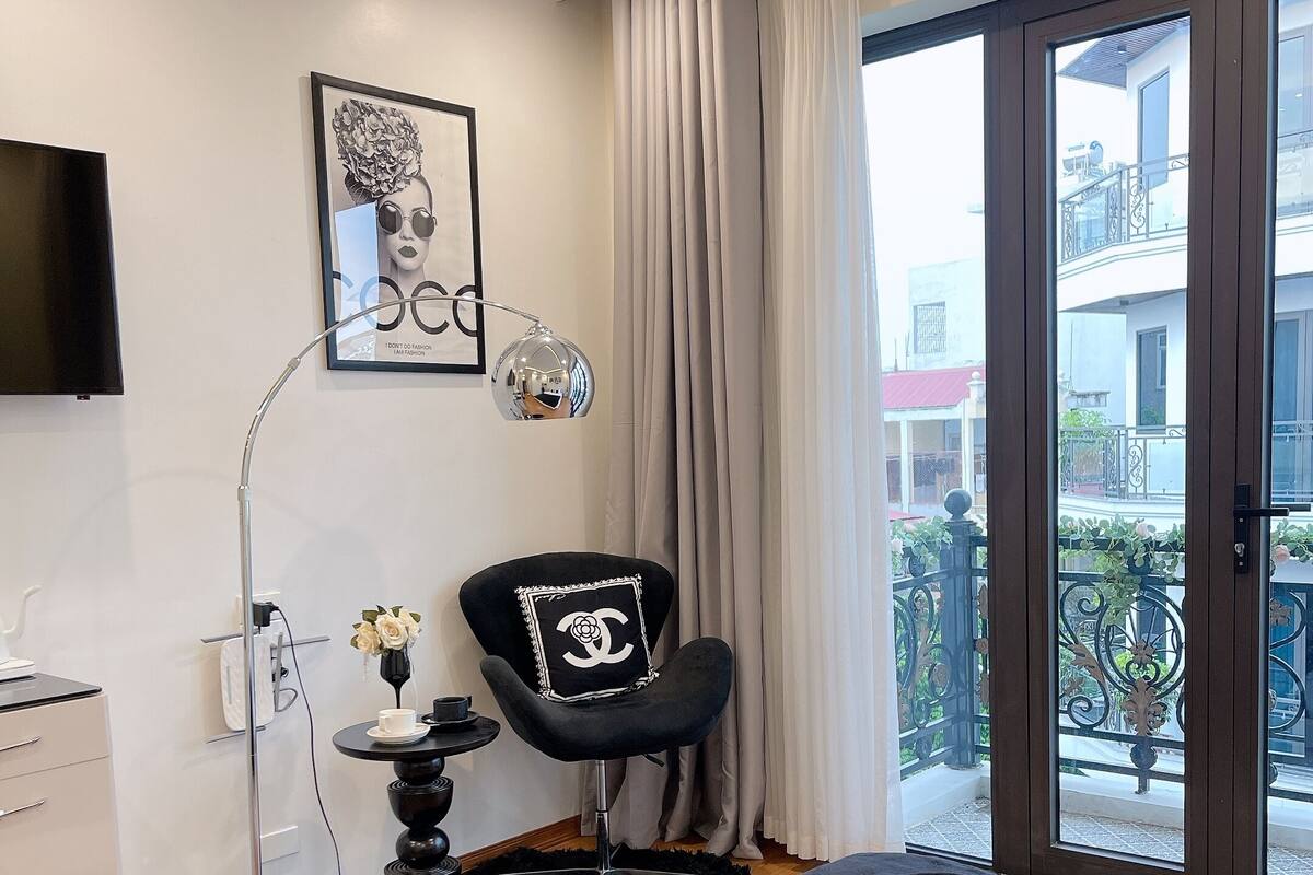 A modern bedroom with a large bed, a black armchair with a Chanel pillow, a standing lamp, and a TV on the wall.