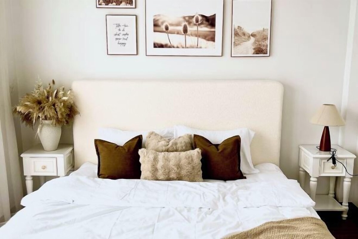 Uma cama bem arrumada com roupa de cama branca e almofadas castanhas, um vaso com flores secas e quadros nas paredes.