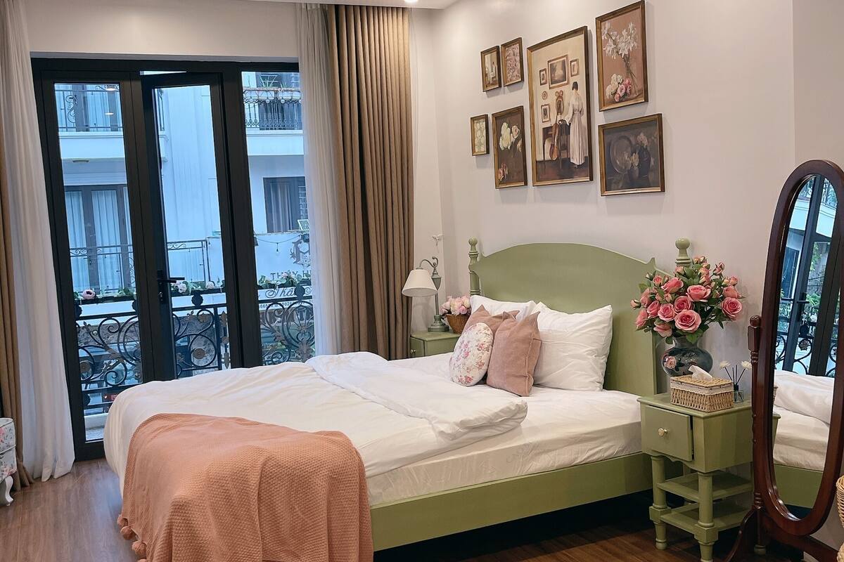 A bedroom with a bed, a nightstand, a mirror, and a window with a view of balconies.