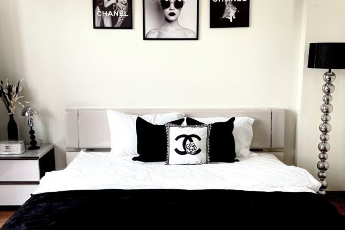 A neatly made bed with white bedding and black and white pillows, including a Chanel logo pillow. There are three framed pictures on the wall above the bed.