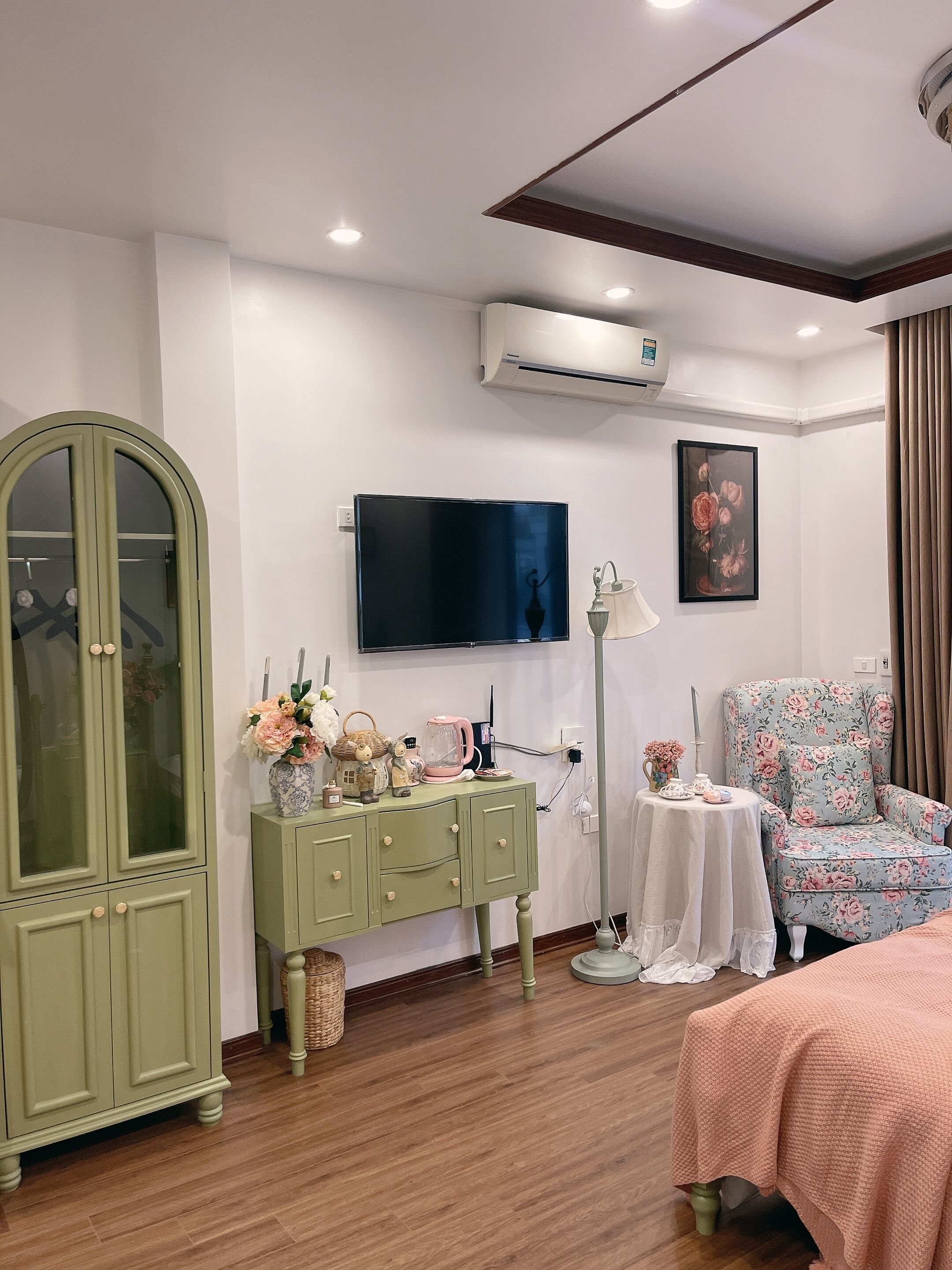 Una habitación con un armario verde, un televisor de pantalla plana montado en la pared, un sillón estampado con motivos florales y una cama con una manta rosa.