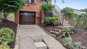 Een tweelaags bakstenen huis met een garage voor de deur, een kleine voortuin met een betonnen pad dat naar de deur leidt, en een metalen trap naar de tweede verdieping.