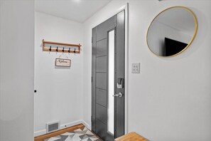 Un pasillo con una puerta de madera oscura, un perchero con un letrero que dice 'Welcome', un espejo redondo con marco dorado, y una alfombra estampada en el suelo.