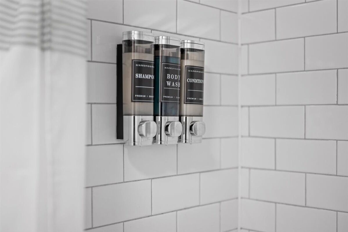 A wall-mounted dispenser with three bottles labeled "Shampoo," "Body Wash," and "Conditioner."