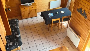 A compact room with a wooden ceiling, a television on a stand, a table with chairs, a radiator, and a window with curtains.