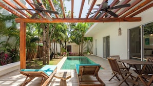 Ein Poolbereich mit Holzliegen, einem Tisch und einer Pergola mit Deckenventilatoren.