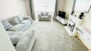 A living room with a grey sofa, a grey armchair, a TV, a fireplace, and a white cabinet. - Stunning Double Bedroom House Closer to the sea (Hartlepool)