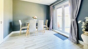 A room with a grey wall, a large window with grey curtains, a wooden floor, and a table with chairs. - Stunning Double Bedroom House Closer to the sea (Hartlepool)