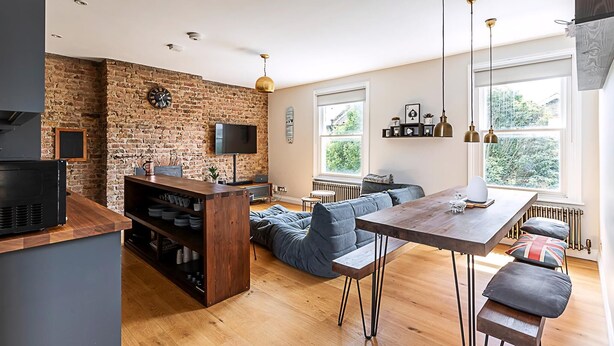 A modern kitchen with a brick wall, wooden flooring, and a dining area with a table and bench. - Rustic and Contemporary Haven in Brixton (London)