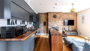 A modern kitchen with a brick wall, a flat-screen TV, and a wooden dining table. - Rustic and Contemporary Haven in Brixton (London)
