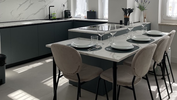 Una cocina moderna con salpicadero de mármol, gabinetes grises y un comedor preparado para seis personas con sillas blancas y una mesa de mármol.