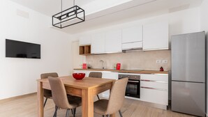 A modern kitchen with white cabinets, a stainless steel refrigerator, a wooden dining table with chairs, a TV mounted on the wall, and a built-in oven.