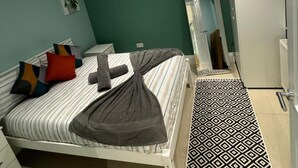 A neatly made bed with striped bedding and a rolled towel, a bedside table with a lamp, a mirror, and a patterned floor.