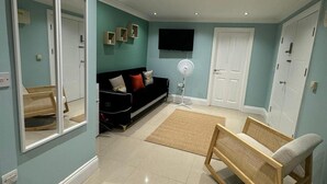 A living room with a black sofa, a wooden bench, a standing fan, and a television on the wall.