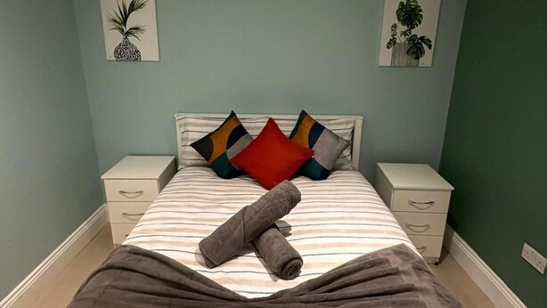 A neatly made bed with striped bedding, multiple pillows, and a rolled towel. There are two bedside tables with drawers, and the walls are painted in a light teal color.