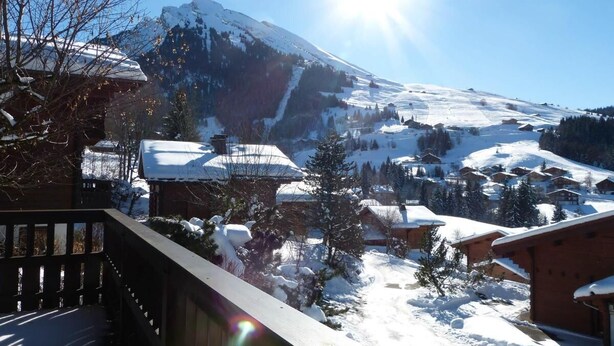 A snow-covered mountain with a clear sky and a sun shining. There are buildings with snow on their roofs and a wooden deck in the foreground.
