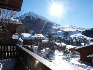A snow-covered mountain with a clear sky and a sun shining. There are buildings with snow on their roofs and a wooden deck in the foreground. - Chalet, balcony, fireplace or stove, tv, ski locker, 102m², La Clusaz (La Clusaz)