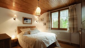 A bedroom with a wooden ceiling, a bed with white bedding, a nightstand with a lamp, a window with curtains, and a radiator.