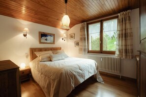 A bedroom with a wooden ceiling, a bed with white bedding, a nightstand with a lamp, a window with curtains, and a radiator.