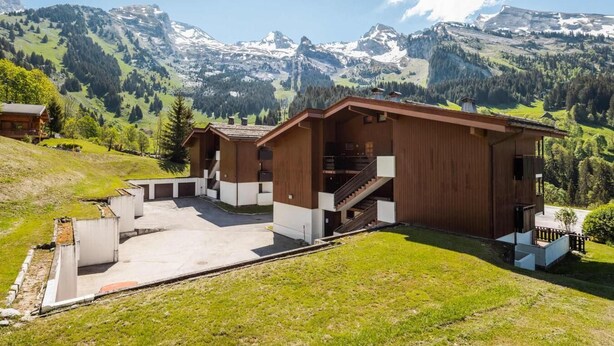 A chalet with a wooden exterior, a covered outdoor area, and a view of snow-capped mountains.