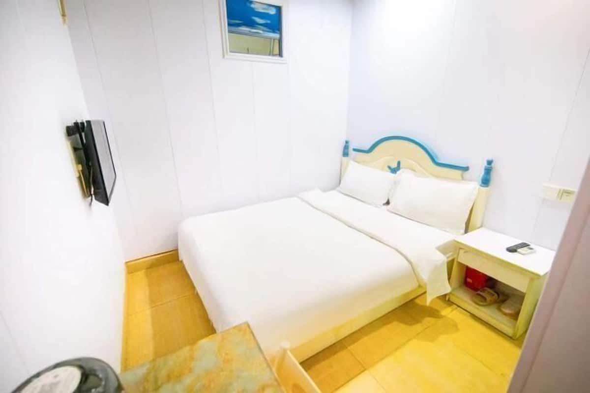 Un lit simple avec une tête de lit en bois, une table de chevet avec un téléphone et une petite étagère avec quelques objets.