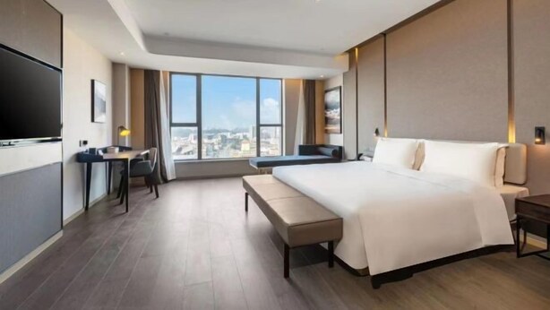 A modern hotel room with a large bed, a desk, a chair, a bench, and a TV.