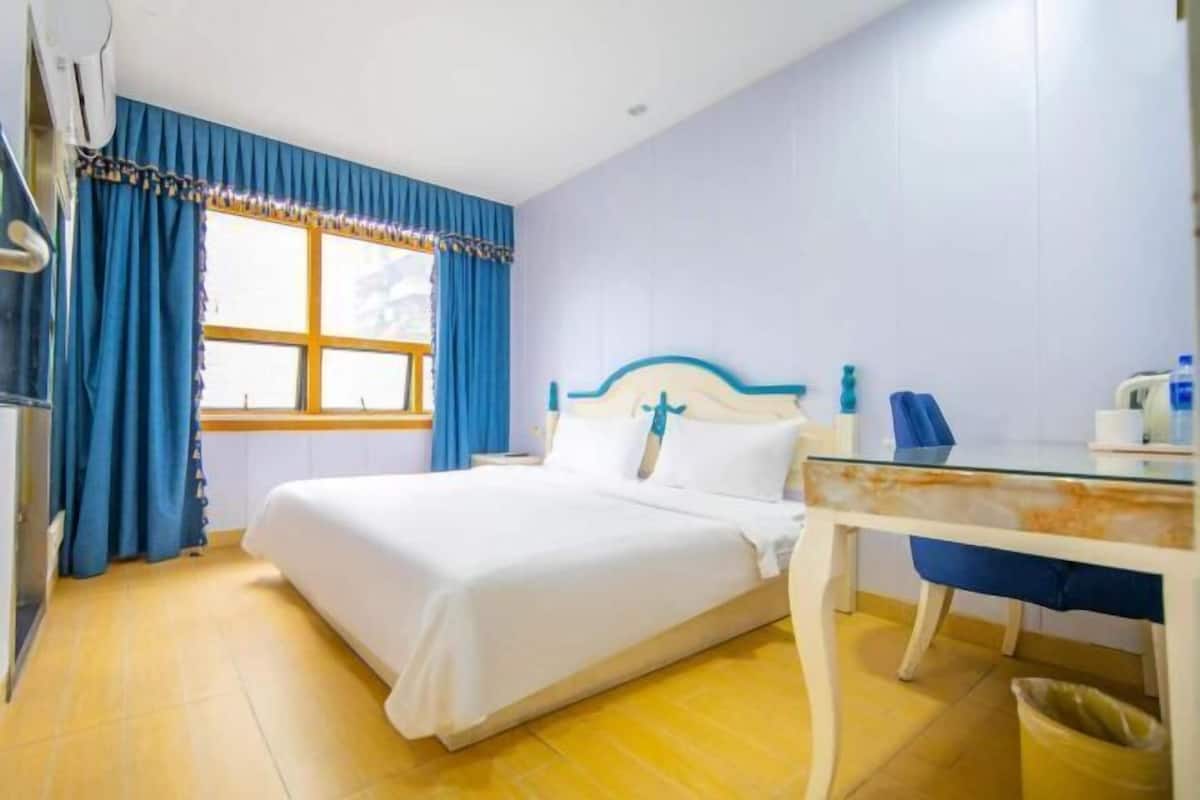 Une chambre d’hôtel comprenant un lit, un bureau avec une chaise, une fenêtre avec des rideaux et une poubelle.