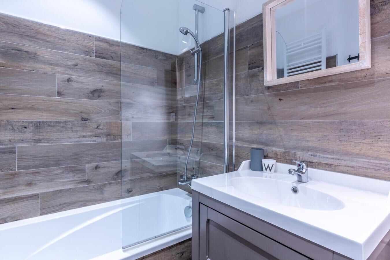 A modern bathroom with a glass shower enclosure, a white bathtub, and a vanity with a mirror and two cups.