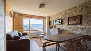 A cozy living room with a sofa, a dining table, and a kitchen area with a stone wall and a TV. - 1st floor, view mountain, balcony, tv, ski locker, 37m², La Clusaz (La Clusaz)