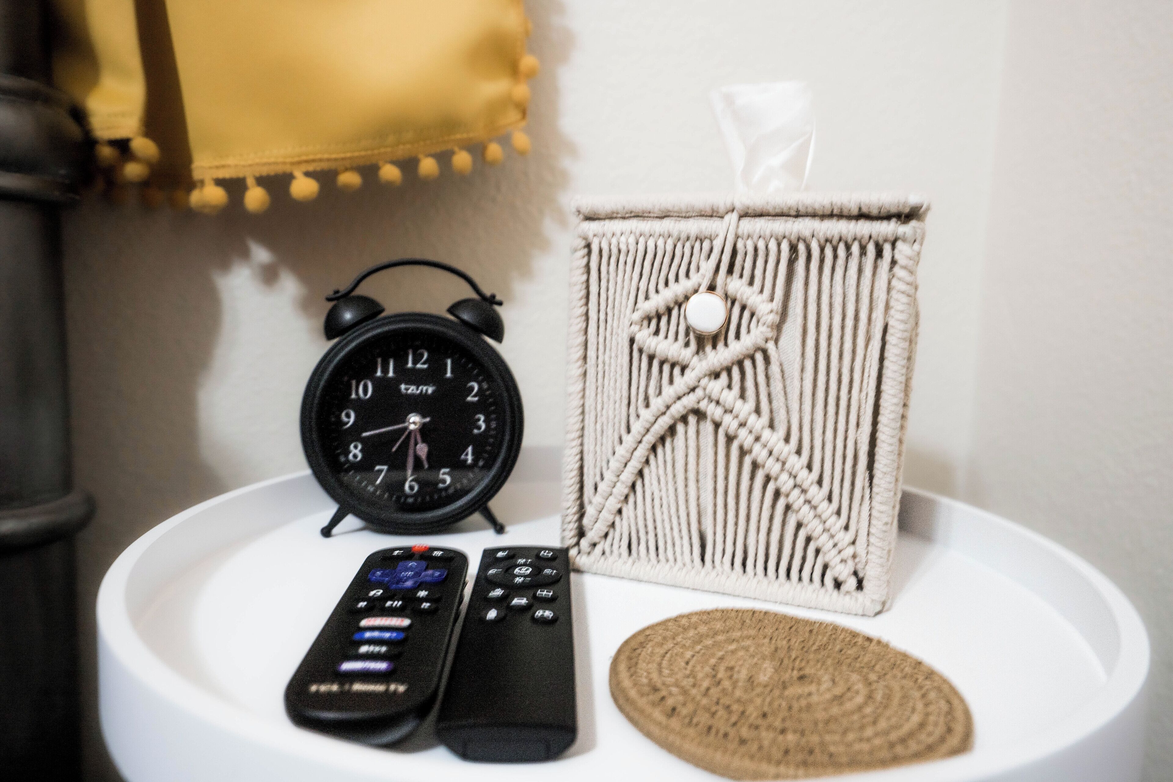 A white round table with a black alarm clock, two remote controls, and a macrame wall hanging.