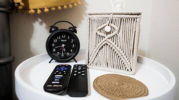 A white round table with a black alarm clock, two remote controls, and a macrame wall hanging.