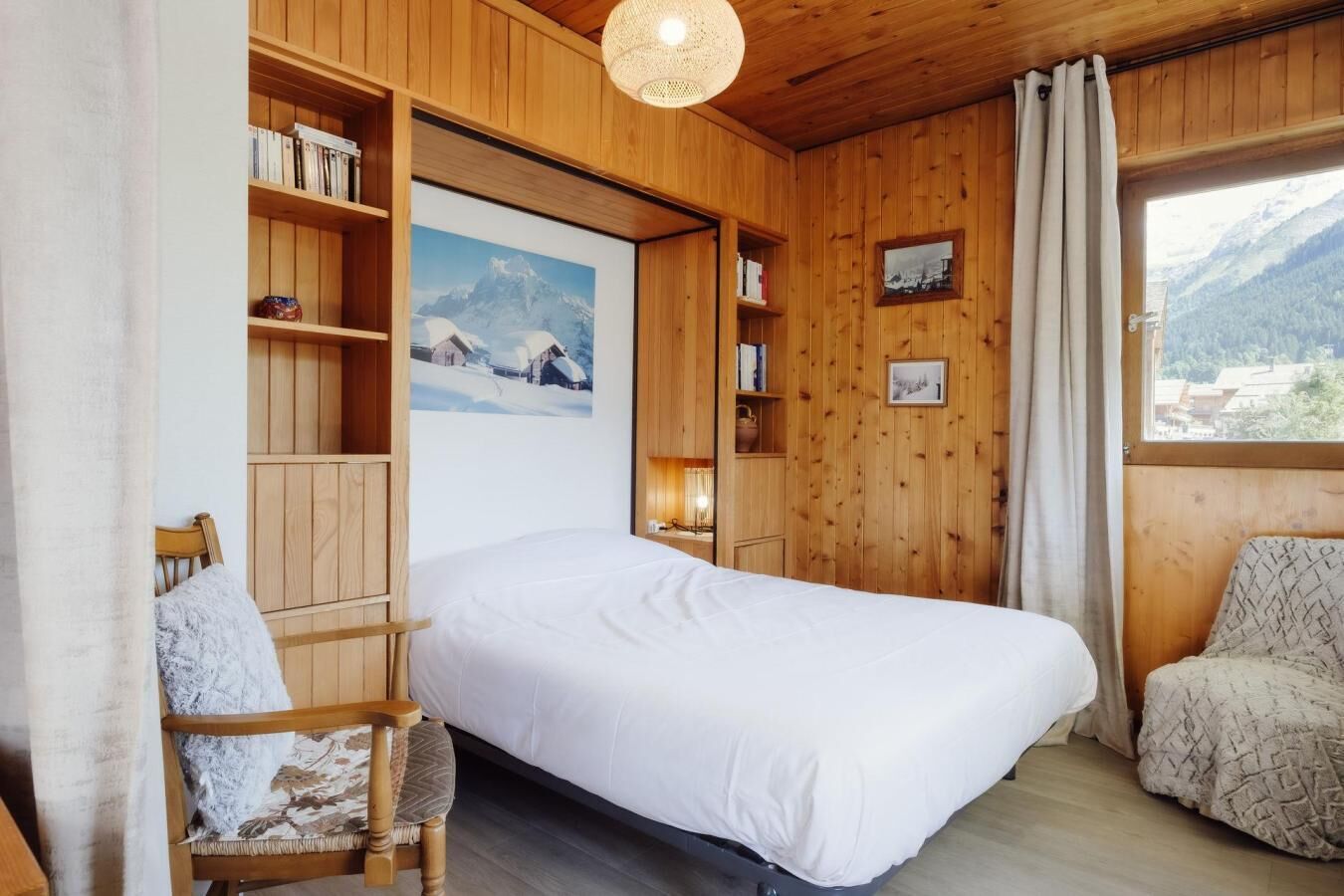 A cozy room with a bed, wooden shelves, a chair, and a window with a mountain view.