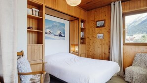A cozy room with a bed, wooden shelves, a chair, and a window with a mountain view.