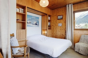 Ein gemütliches Zimmer mit einem Bett, Holzböden, einem Sessel und einem Fenster mit Blick auf die Berge.