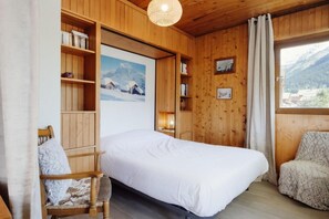 A cozy room with a bed, wooden shelves, a chair, and a window with a mountain view.