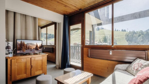 A cozy room with a wooden ceiling, a large window with a view of a green landscape, a wooden cabinet with a television, and a sofa with cushions.