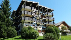 A multi-story building with balconies, surrounded by greenery and a clear sky.