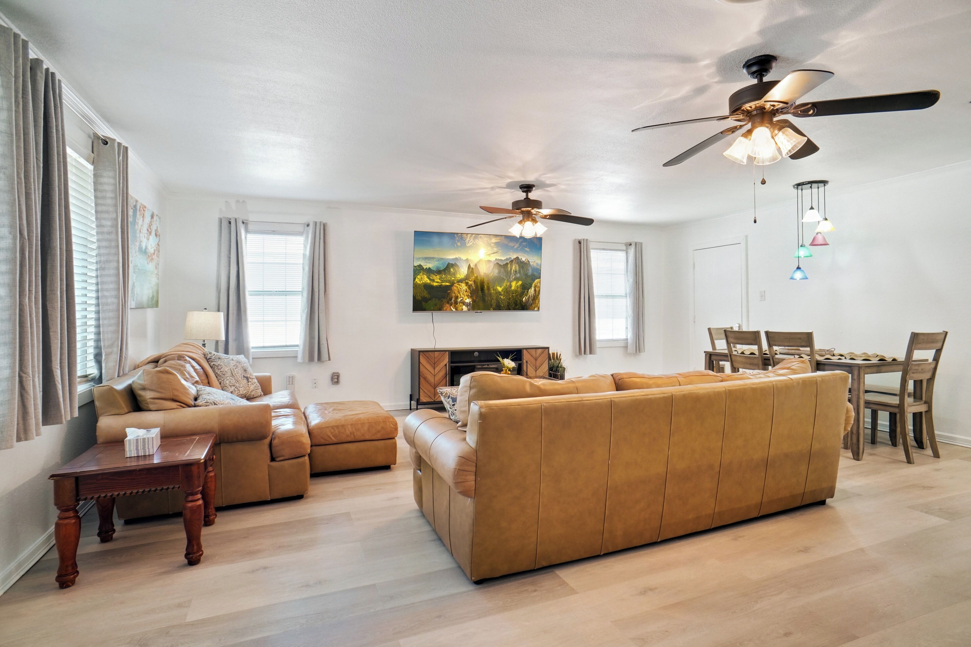 A living room with a sofa, ottoman, wooden coffee table, and a painting on the wall. There is a ceiling fan and pendant lights.