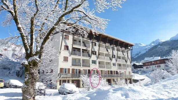 A multi-story building with balconies, surrounded by snow-covered trees and mountains in the background.