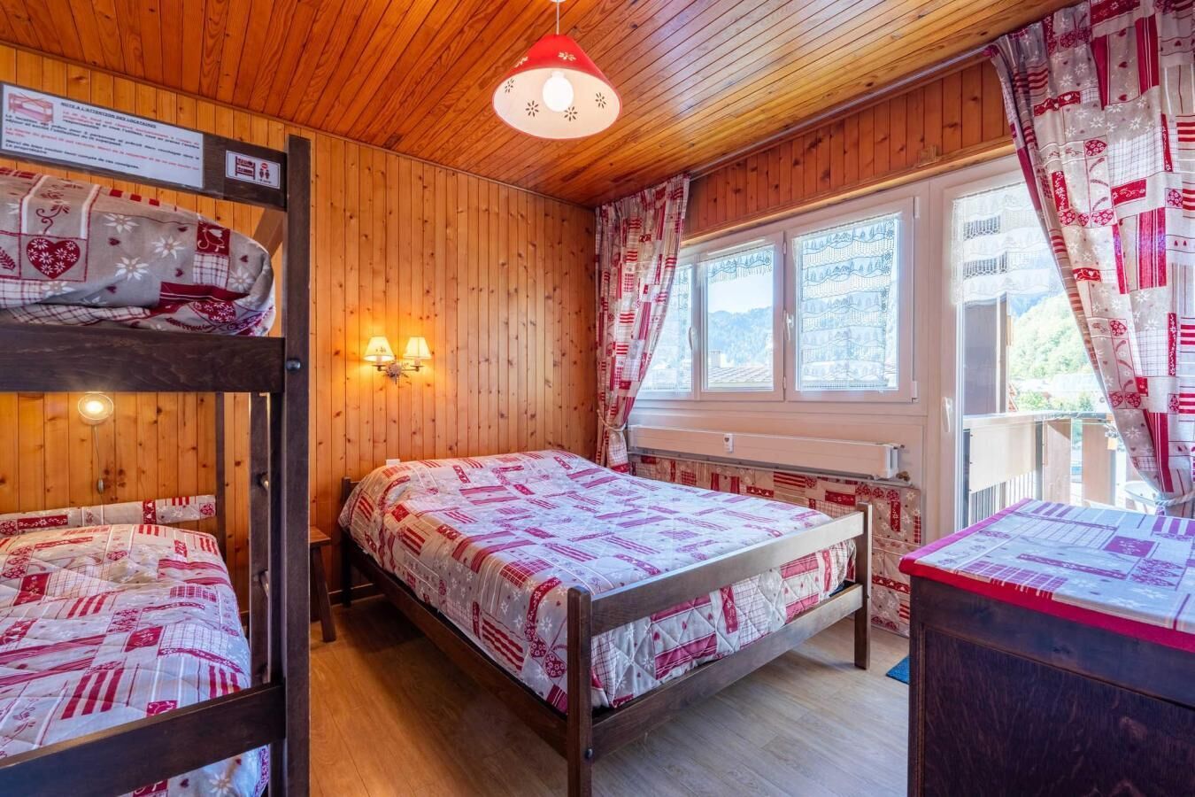 Una habitación acogedora con paredes de madera y una cama litera. Hay dos mesitas de noche con lámparas, una ventana con cortinas y vistas al exterior.