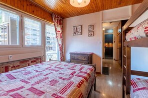 A bedroom with a wooden ceiling, a bed with a quilt, a dresser, and a view of the city through the window.