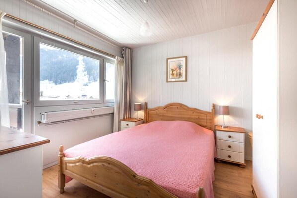 A bedroom with a wooden bed, two bedside tables with lamps, a window with curtains, and a framed picture on the wall.