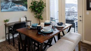 Uma sala de jantar com mesa de madeira posta para quatro pessoas, um vaso de planta e uma pintura de paisagem de montanha.