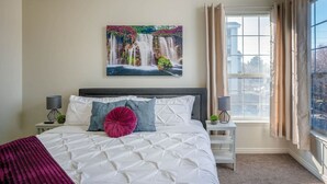 Quarto com uma cama grande, janela com cortinas, mesa de cabeceira com abajur e pintura pendurada na parede retratando uma cachoeira.