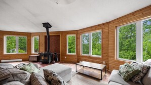A cozy cabin interior with a central wood-burning stove, large windows offering views of greenery, and a comfortable seating area with cushions. - Zulu Zen waterfront cottage (Blue Sea)