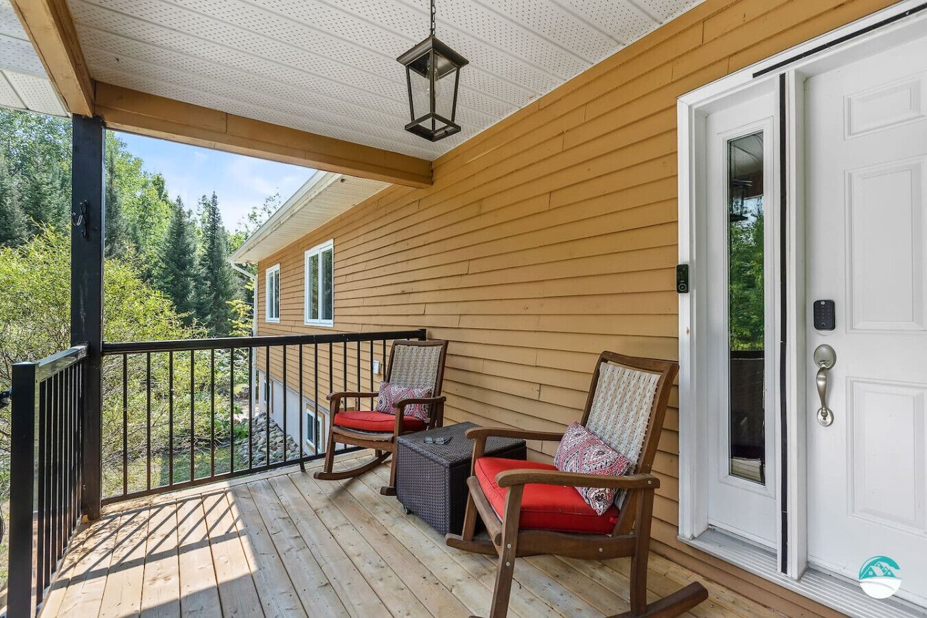 A wooden deck with two rocking chairs and a table, a white door with a keypad lock, and a view of trees and a house in the background.