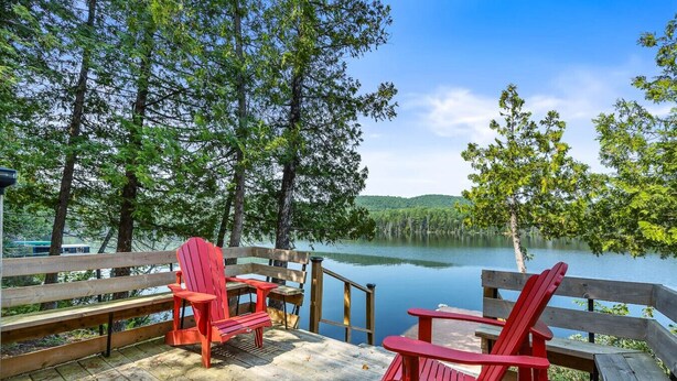 A wooden deck with two red Adirondack chairs overlooking a calm lake surrounded by trees. - Zulu Zen waterfront cottage (Blue Sea)