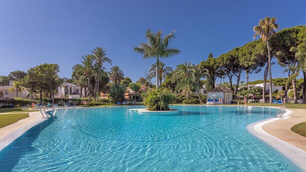 A large, clear swimming pool surrounded by palm trees and lounge chairs, with a small island in the center. - 3 Bedroom, 3 Bathroom Beachside Apartment (Estepona)
